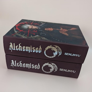 May include: Two stacked hardback books with the title "Alchemised" in a gothic font. The covers are dark burgundy with a detailed illustration and the author's name, "Senlinyu". The top book's cover features a dark illustration with a red circular design.