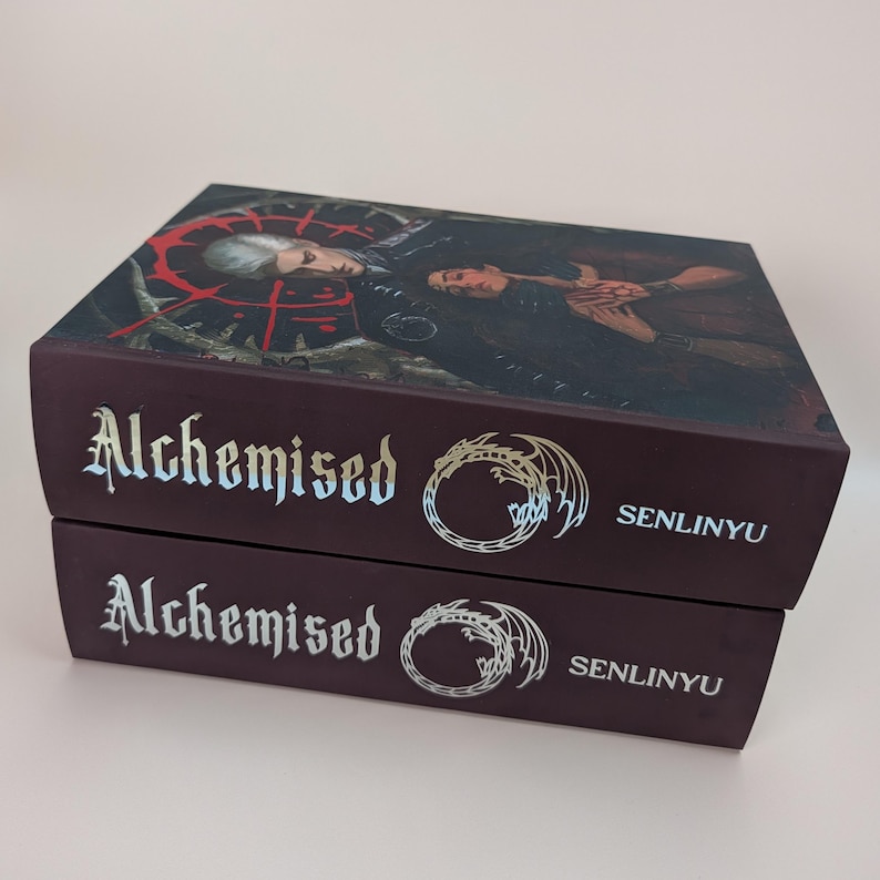 May include: Two stacked hardback books with the title "Alchemised" in a gothic font. The covers are dark burgundy with a detailed illustration and the author's name, "Senlinyu". The top book's cover features a dark illustration with a red circular design.