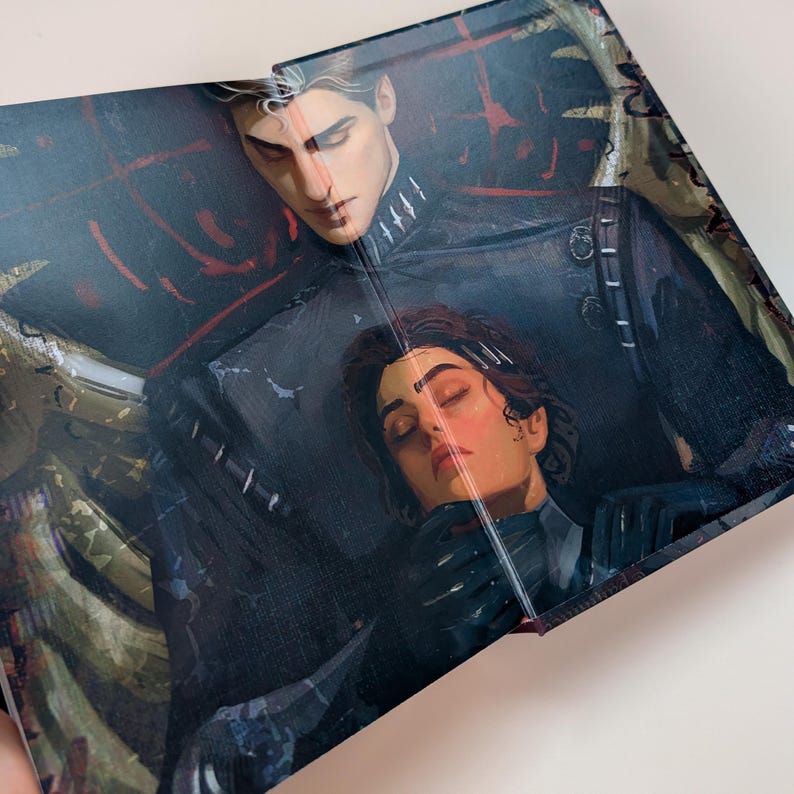 May include: A book cover featuring a detailed illustration of two figures. One figure, with light skin and dark hair, is embracing another figure with dark hair and closed eyes. The artwork has a dark color palette with hints of red and gold.
