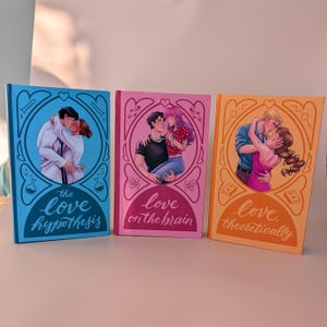 Puede incluir: Tres libros de tapa dura con ilustraciones románticas. Los libros son de color azul, rosa y naranja. Cada libro presenta una pareja abrazándose, con títulos como "the love hypothesis" y "love on the brain". Los libros se exhiben sobre una superficie clara.