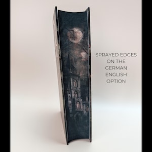 May include: A hardcover book with a gothic castle illustration under a full moon. The book's cover is dark with a detailed image of a castle, trees, and a cloudy sky. The text on the side reads "Sprayed edges on the German English option."