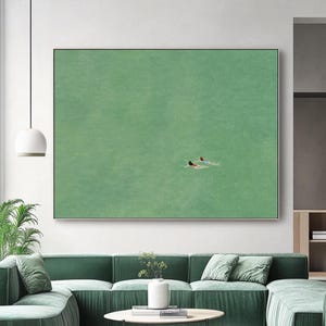 Könnte beinhalten: Ein großes rechteckiges Wandbild mit einem einfarbigen salbeigrünen Hintergrund und zwei kleinen Figuren, die in der Mitte schwimmen. Das Gemälde ist in einem dünnen Metallrahmen gefasst und hängt an einer weißen Wand über einem grünen Samtsofa.