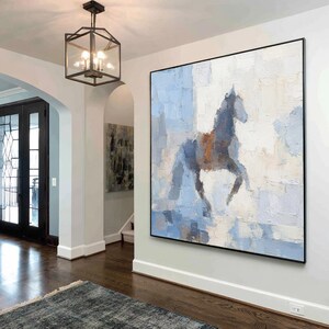 May include: Abstract painting of a horse in blue, white, and brown tones. The artwork is displayed in a modern interior, mounted on a white wall with a black frame. The painting's style is impressionistic. The room has a dark wooden floor.