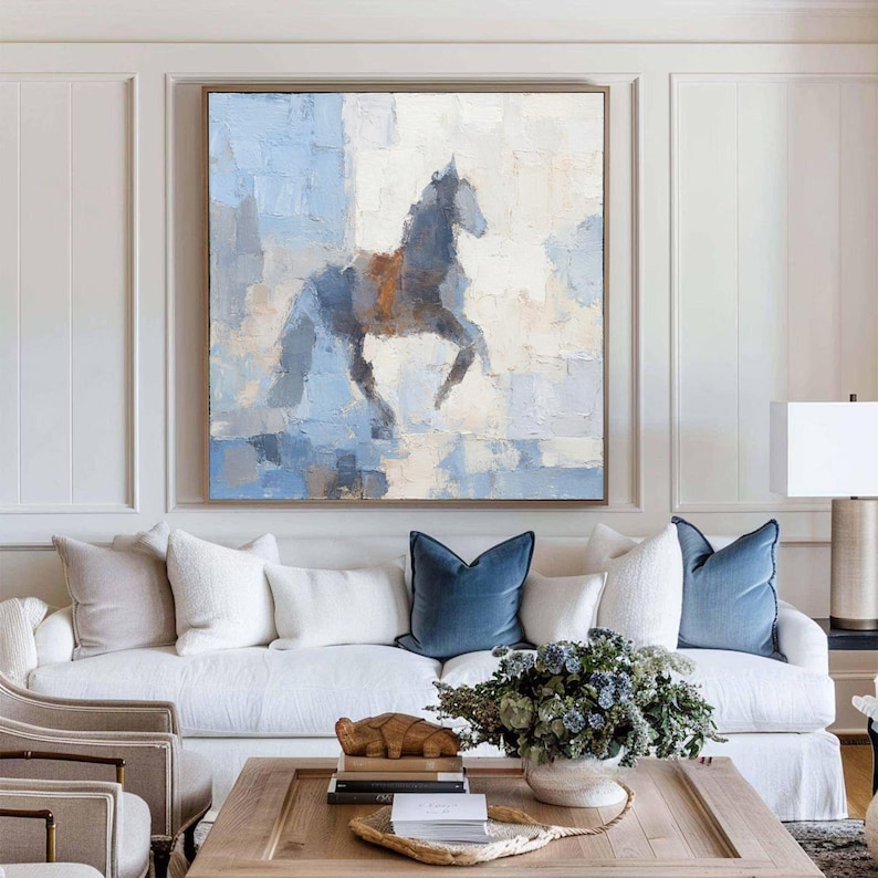 May include: Large framed abstract painting of a horse in shades of blue, grey, and brown. The artwork is displayed above a white sofa with blue velvet throw cushions. The room has a neutral colour palette with a wooden coffee table.