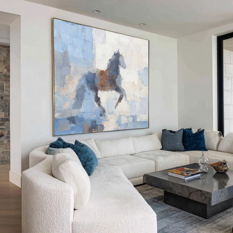May include: Large abstract painting of a rearing horse in shades of blue, grey, and brown. The artwork is displayed on a white wall above a white sectional sofa with blue cushions. The painting has a gold frame.