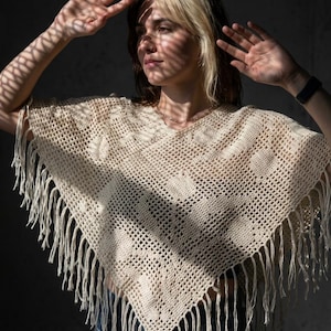 May include: A cream-coloured crochet poncho featuring an open-knit grid pattern with integrated diamond shapes and long, dangling fringe along the bottom edges. The garment has a V-neckline and a triangular silhouette that drapes over the shoulders.