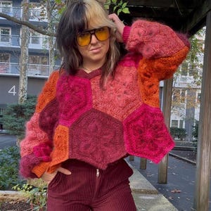May include: A colorful, patchwork crochet sweater in shades of red, pink, and orange. The sweater features a geometric design with hexagonal panels. The sleeves are long and flared. The person is wearing burgundy corduroy pants and yellow-tinted glasses.