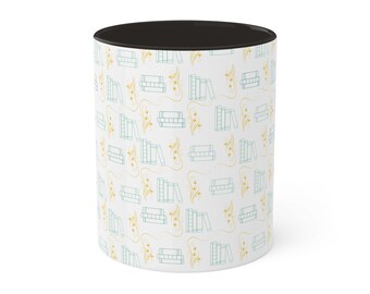 May include: White ceramic mug with a black rim and a pattern of yellow flowers and stacks of blue books.