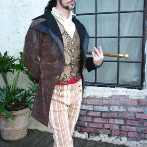 May include: A man wearing a brown jacket with a black velvet collar, a gold and brown patterned waistcoat, a red sash, and striped trousers. He is wearing a black top hat with a gold embellishment and holding a wooden walking stick with a gold knob.