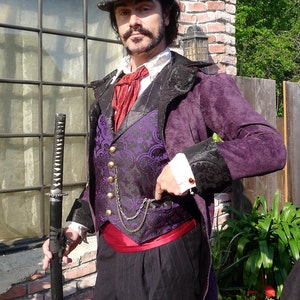 Puede incluir: Un hombre con una chaqueta de terciopelo morado con un chaleco estampado en negro y morado, una corbata de lazo roja y un sombrero de copa negro. Está sosteniendo una espada katana negra.