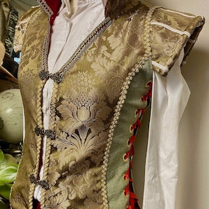 May include: A gold brocade vest with a red and gold collar, green and gold side panels, and a red ribbon lacing up the back. The vest has a floral pattern and decorative metal buttons.