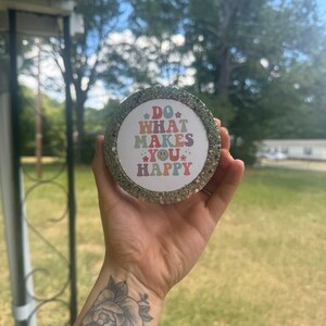 May include: A round, glittery, gold-colored coaster with a white background and the text "DO WHAT MAKES YOU HAPPY" in rainbow colors. The text is surrounded by small flowers and a smiley face.