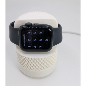 May include: A black smartwatch with a passcode screen rests on a white charging stand. The watch features a black strap and a digital display showing numbers. The charging stand has a textured design.