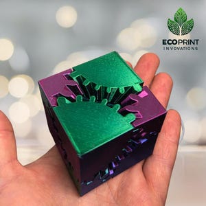 May include: A 3D printed cube with interlocking gears, featuring a green and purple iridescent finish. The cube is held in a hand, with the text "ECOPRINT INNOVATIONS" visible in the background.