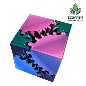 Puede incluir: Un cubo impreso en 3D con engranajes entrelazados. El cubo está hecho de tres colores: rosa, verde y azul. El cubo es un diseño único e intrincado.