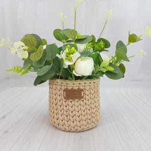 May include: A beige crocheted basket filled with white flowers and green foliage. The basket has a small brown tag that reads "Big Hook Yarn". The arrangement includes white peonies, eucalyptus, and other greenery, creating a natural, decorative display.