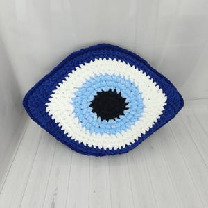 May include: A handmade crochet pillow shaped like an eye, featuring concentric layers of yarn in dark blue, white, light blue, and a solid black center. The textured, soft-looking textile construction forms a distinct almond-shaped design.
