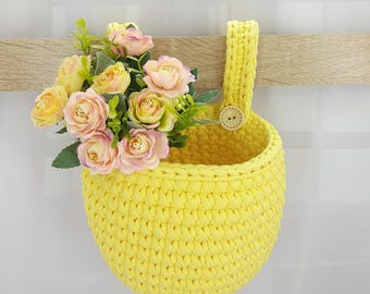 May include: A bright yellow crocheted hanging basket with a single loop handle and a small wooden button detail. The basket is filled with a cluster of artificial pink and yellow roses with green leaves, creating a vibrant floral arrangement.
