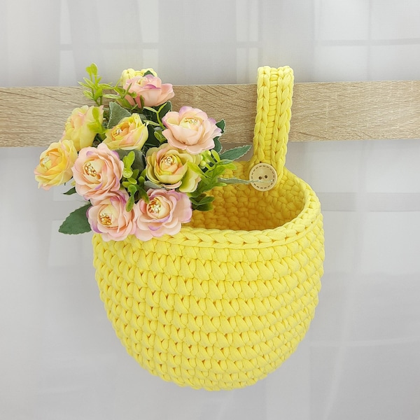 Könnte beinhalten: Ein hellgelber, gehäkelter Hängekorb mit einer einzelnen Schlaufe als Griff und einem kleinen Holzknopf. Der Korb ist mit einem Strauß aus künstlichen rosa und gelben Rosen sowie grünen Blättern gefüllt, was ein lebendiges Blumenarrangement ergibt.