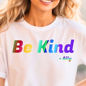 May include: White t-shirt with the words "Be Kind" in a rainbow gradient font. The text "+ Ally" is below the main text, also in a rainbow gradient. The shirt has short sleeves.