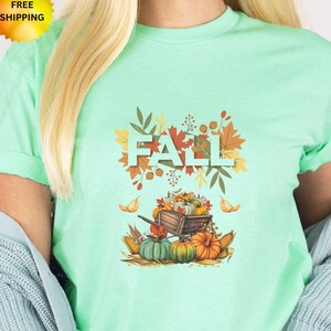 May include: A mint green short-sleeve t-shirt featuring a graphic print of a wooden cart filled with orange and green pumpkins, surrounded by autumn leaves and the word FALL in large block letters centered above the cart illustration.