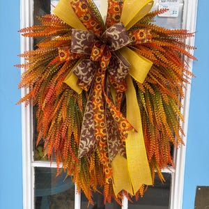 May include: A teardrop-shaped autumn wreath featuring dense, feathery foliage in shades of orange, yellow, and green. A large, multi-layered decorative bow sits at the top, crafted from yellow ribbon and brown ribbon with sunflower and swirl patterns.