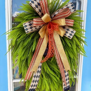 May include: A teardrop-shaped wreath made of bright green fern foliage, adorned with a large decorative bow. The bow features layers of ribbons with a blue and white diamond pattern, a red wavy line design, and solid gold glittery fabric.