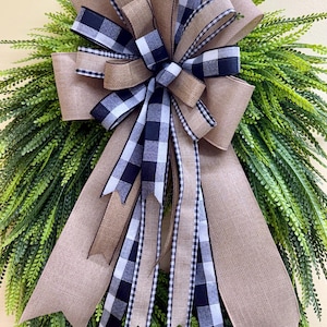May include: A decorative wreath featuring a large, multi-layered bow made of tan burlap and black and white buffalo check ribbon. The bow is centered over a dense arrangement of long, pointed green fern-like foliage with a textured appearance.