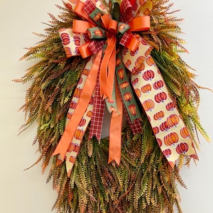 May include: A teardrop-shaped wreath featuring dense, textured foliage in shades of green, orange, and brown. A large, multi-layered bow sits at the top, crafted from ribbons with pumpkin patterns, solid orange, and dark green plaid designs.