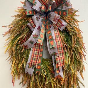 May include: A teardrop-shaped wreath made of green, yellow, and orange fern-like foliage. A large decorative bow sits at the top, featuring ribbons with patterns of orange pumpkins, autumn maple leaves, and a multi-colored plaid design.