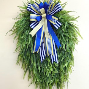 May include: A green artificial wreath with a blue, yellow, and white ribbon bow. The bow is tied with a large knot and has long tails.