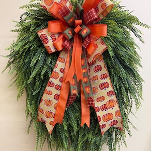 May include: A fall-themed wreath with a lush green base and a large bow. The bow features ribbons with pumpkin patterns, orange solid ribbons, and a plaid pattern. The wreath is perfect for autumn decor.