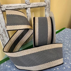 May include: A roll of decorative ribbon with a black and white polka dot pattern. The ribbon has a natural burlap texture with black trim. The ribbon is displayed in front of a distressed white wooden window frame.