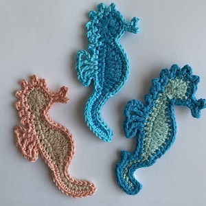 Pode incluir: Três ornamentos de cavalo-marinho de crochê em diferentes cores: azul claro, azul-turquesa e pêssego. Os cavalos-marinhos estão todos virados para o lado direito da imagem.