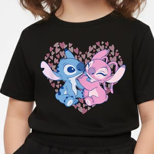 以下が含まれることがあります： 黒いTシャツ。ハート型のデザインで、漫画のキャラクターが描かれています。青いキャラクターとピンクのキャラクターが抱き合っており、小さなピンクのハートの背景があります。