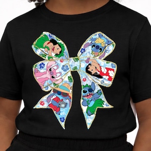 May include: Black t-shirt with a large bow design showcasing cartoon characters. The bow has a blue floral pattern with gold edging. The characters are in various poses and outfits, adding a playful touch to the shirt.