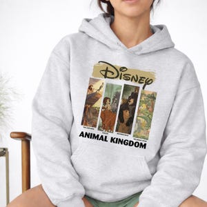 Peut inclure: Un sweat &agrave; capuche gris clair avec un motif Disney. Le motif comprend le logo Disney en or et quatre panneaux rectangulaires avec des images de "Le Roi Lion", "Tarzan", "Fr&egrave;re des ours" et "Le Livre de la Jungle". Les mots "ANIMAL KINGDOM" sont imprim&eacute;s en dessous.