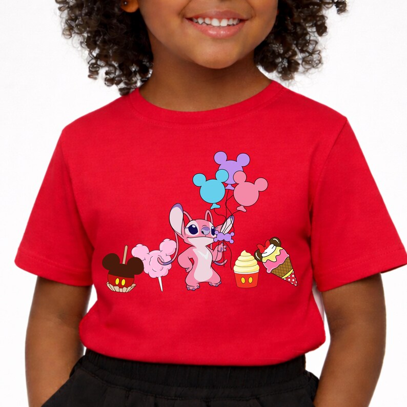 Op de afbeelding: Een rood t-shirt met een stripfiguur die ballonnen vasthoudt en omringd is door snoep. De afbeelding bevat een Mickey Mouse-vormige appel, suikerspin, een cupcake en een ijshoorntje. Het shirt wordt door een kind gedragen.