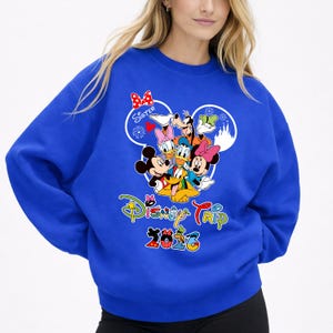 May include: Royal blue sweatshirt with a Disney Trip 2023 graphic. The design features Mickey Mouse, Minnie Mouse, Donald Duck, Goofy, and Daisy Duck. The word "Sister" is above the characters in a heart-shaped design.