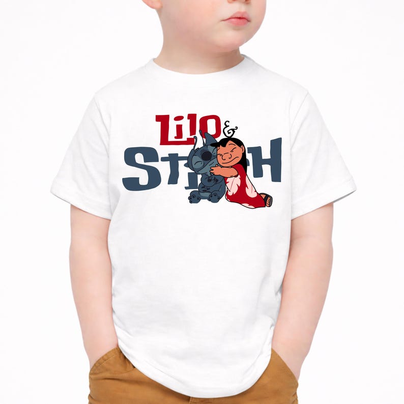K&ouml;nnte beinhalten: Ein wei&szlig;es kurz&auml;rmeliges T-Shirt mit einer Grafik der Figuren Lilo und Stitch, die sich umarmen. Der Text Lilo & Stitch ist in roter und dunkelblauer Schrift &uuml;ber und hinter den Figuren aufgedruckt, die in ihren klassischen Farben dargestellt sind.