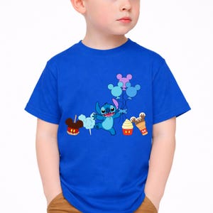 Op de afbeelding: Een blauw t-shirt met een cartoon Stitch-personage dat ballonnen en lekkernijen vasthoudt. De afbeelding bevat een Mickey Mouse-vormige snoepappel, suikerspin, een cupcake en een ijshoorntje. Het shirt wordt door een kind gedragen.