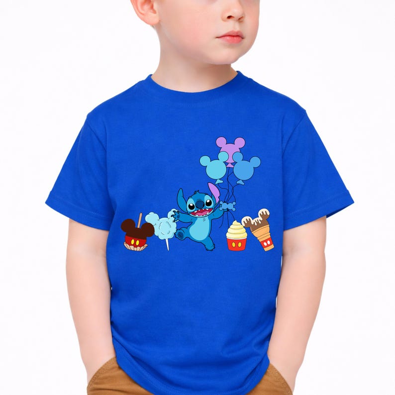 Op de afbeelding: Een blauw t-shirt met een cartoon Stitch-personage dat ballonnen en lekkernijen vasthoudt. De afbeelding bevat een Mickey Mouse-vormige snoepappel, suikerspin, een cupcake en een ijshoorntje. Het shirt wordt door een kind gedragen.