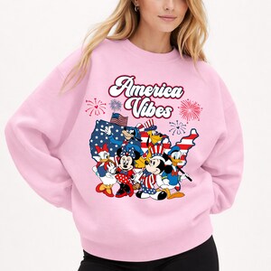 May include: A light pink sweatshirt featuring a graphic of Disney characters in patriotic attire. The design includes Mickey Mouse, Minnie Mouse, Donald Duck, and Daisy Duck, along with the text "America Vibes" and fireworks.