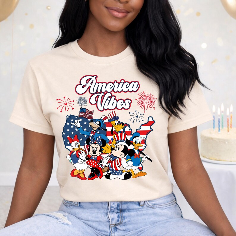 May include: Cream-colored t-shirt featuring a graphic of cartoon characters in patriotic attire. The design includes the text "America Vibes" above a map of the United States, with fireworks and stars.