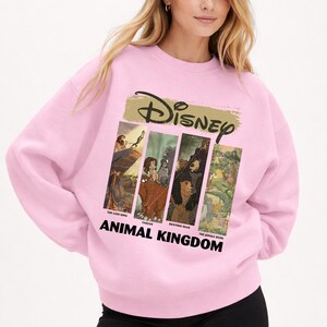 Peut inclure: Un sweat-shirt rose clair avec le logo Disney et des images de "Le Roi Lion", "Tarzan", "Fr&egrave;re des ours" et "Le Livre de la Jungle". Les mots "ANIMAL KINGDOM" sont imprim&eacute;s en gras, en noir, en dessous.