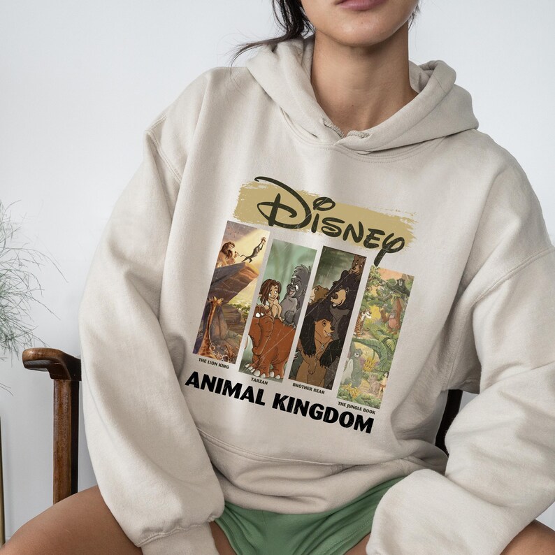 Peut inclure: Sweat &agrave; capuche beige avec un motif Disney repr&eacute;sentant des sc&egrave;nes du Roi Lion, Tarzan, Fr&egrave;re des ours et Le Livre de la Jungle. Le mot "Disney" est au-dessus des images, et "Animal Kingdom" est imprim&eacute; en dessous.