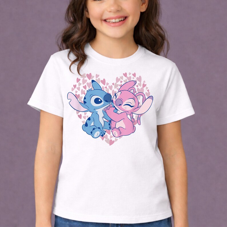 以下が含まれることがあります： 白いTシャツ。青とピンクの2人のキャラクターが、ピンクのハートのハート型のデザインの中で抱き合っている漫画のイラストが描かれています。キャラクターはアニメ映画リロ＆スティッチからです。