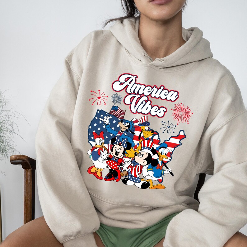 May include: Beige hoodie featuring a graphic of cartoon characters in patriotic attire, including Mickey and Minnie Mouse, Donald and Daisy Duck, and Goofy, with the text "America Vibes" and fireworks.