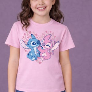 以下が含まれることがあります： ピンク色のTシャツ。リロ＆スティッチのキャラクター、スティッチとエンジェルのグラフィックが描かれています。青いスティッチとピンクのエンジェルが、小さなピンクのハートでできたハートの中で抱き合っています。半袖のシャツです。