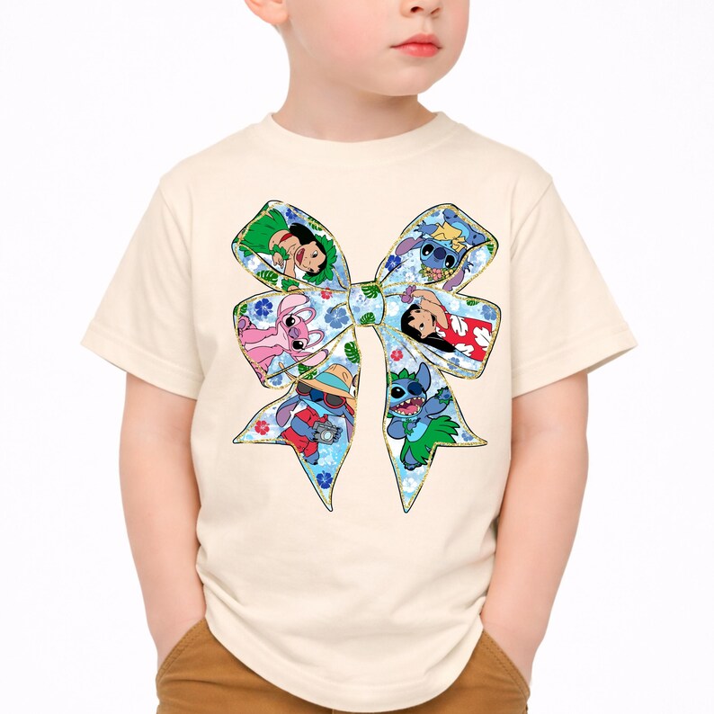 May include: A cream-coloured t-shirt featuring a large bow design with characters from the animated film Lilo & Stitch. The bow is decorated with a floral pattern and images of the characters in various poses.